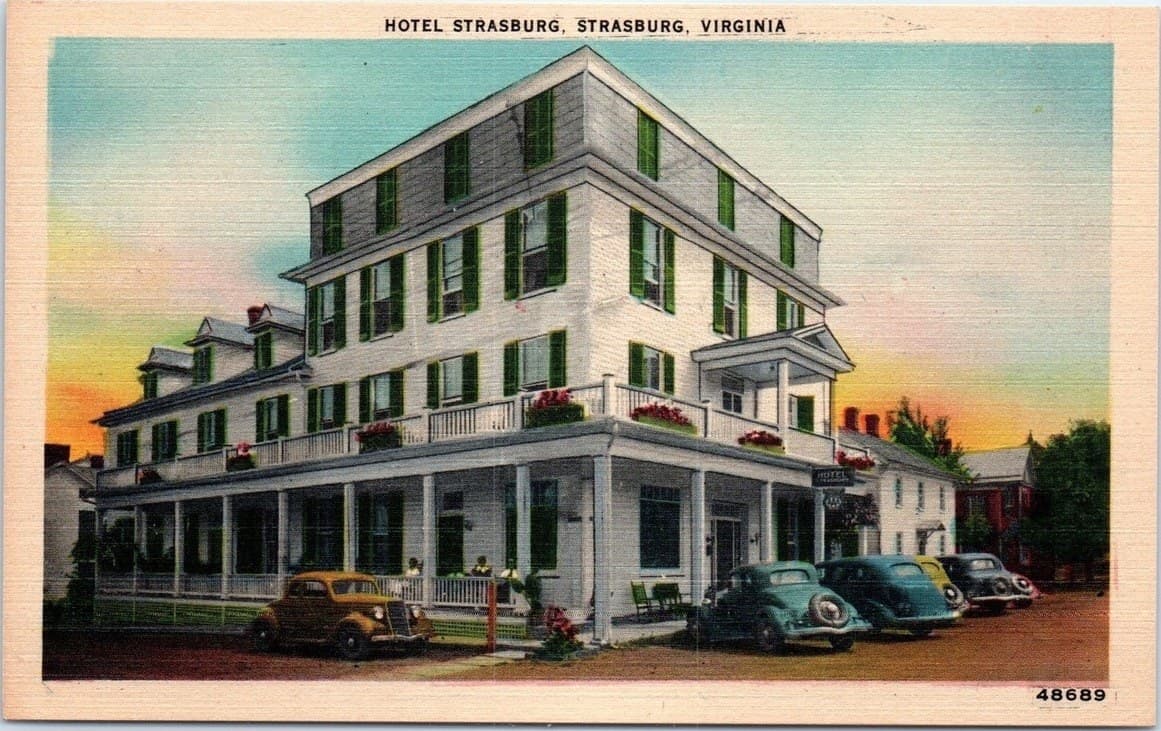 A colour-tinted linen postcard captioned Hotel Strasburg, Strasburg, Virginia. A three-storey white clapboard hotel with green shutters, a wraparound porch and window boxes of red flowers, with four late-1930s cars parked alongside under a pastel sunset sky.