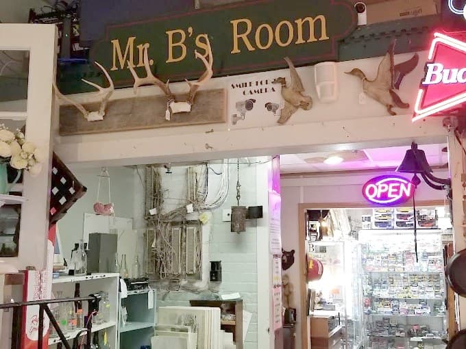 Inside the Strasburg Emporium. A carved wooden sign reading Mr B's Room hangs above a doorway, with mounted antlers, a wooden goose and a small bell beside it. A neon Open sign glows over glass display cases in the room beyond.